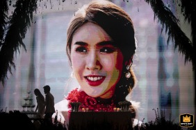 At the Four Seasons Bangkok, the bride’s joyful face beams from a massive digital screen while the newlywed couple, silhouetted and small in comparison, walk together past the vibrant display.